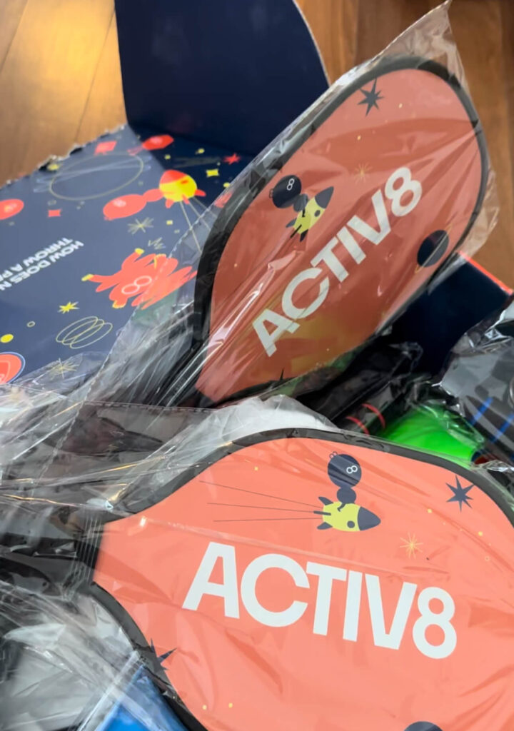 Two pickle ball paddles with the Activ8 logo are in focus, one layered over the other. They're partially wrapped in plastic and have a cartoonish design with stars and a character on the orange surface. In the background, there's an open box with a similar playful design and more equipment partially visible, suggesting the unpacking of an Activ8 active play kit.